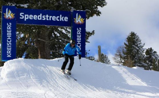 Ein Skifahrer in einer blauen Jacke fährt eine verschneite Piste unter einem Schild mit der Aufschrift "Speedstrecke Kreischberg" hinunter, umgeben von Bäumen.