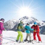 Vier Personen in Skiausrüstung stehen auf einem verschneiten Berghang mit Skiliften und sonnenbeschienenen Bergen im Hintergrund.