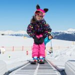 Zwei Kinder in farbenfrohen Skikostümen fahren auf einem Förderband über einen verschneiten Berghang, mit klarem blauen Himmel und fernen Bergen im Hintergrund.