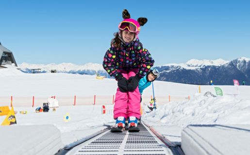 Zwei Kinder in farbenfrohen Skikostümen fahren auf einem Förderband über einen verschneiten Berghang, mit klarem blauen Himmel und fernen Bergen im Hintergrund.