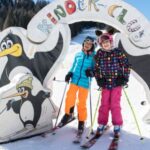 Zwei Kinder in bunten Winterklamotten stehen auf Skiern unter einem mit Pinguinen verzierten Torbogen auf einer verschneiten Skipiste.