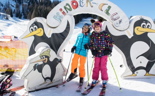 Zwei Kinder in bunten Winterklamotten stehen auf Skiern unter einem mit Pinguinen verzierten Torbogen auf einer verschneiten Skipiste.