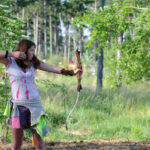 Eine Person zielt mit Pfeil und Bogen im Freien, während eine andere Person in der Nähe steht, mit Bäumen und Grünzeug im Hintergrund.