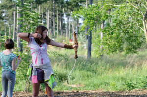 Eine Person zielt mit Pfeil und Bogen im Freien, während eine andere Person in der Nähe steht, mit Bäumen und Grünzeug im Hintergrund.