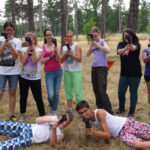 Eine Gruppe junger Leute posiert im Freien in einem Waldgebiet und hält Spielzeugwaffen oder Nachbildungen von Waffen hoch, von denen zwei vor ihnen im Gras liegen und die anderen hinter ihnen stehen.