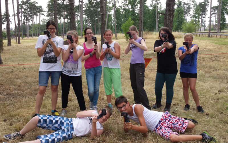 Eine Gruppe junger Leute posiert im Freien in einem Waldgebiet und hält Spielzeugwaffen oder Nachbildungen von Waffen hoch, von denen zwei vor ihnen im Gras liegen und die anderen hinter ihnen stehen.