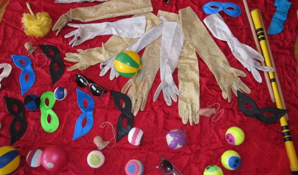 Verschiedene Kostümrequisiten, darunter goldene und silberne Handschuhe, bunte Masken, Bälle, Sonnenbrillen und gelbe Schlagstöcke, sind auf einem roten Stoffhintergrund angeordnet.
