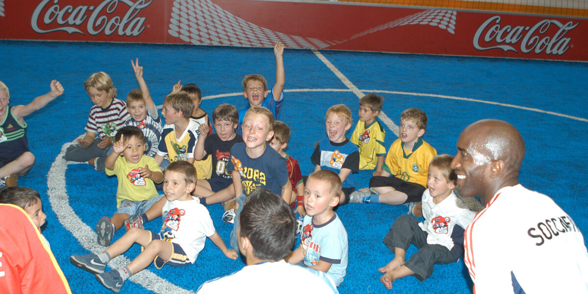 Eine Gruppe von Kindern sitzt auf einem blauen Hallenfußballfeld, lächelt und jubelt, während zwei Erwachsene in "SOCCAFIVE"-Shirts daneben knien.