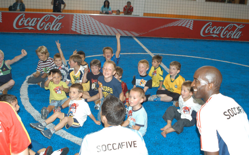 Eine Gruppe von Kindern sitzt auf einem blauen Hallenfußballfeld, lächelt und jubelt, während zwei Erwachsene in "SOCCAFIVE"-Shirts daneben knien.