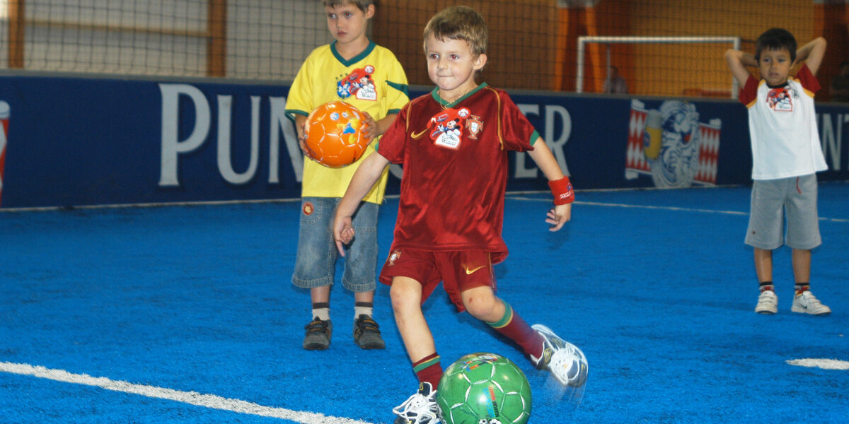 Drei Jungen stehen auf einem blauen Hallenfußballfeld, einer davon in einem roten Trikot, der einen grünen Fußball kickt, während die anderen zusehen.