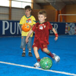 Drei Jungen stehen auf einem blauen Hallenfußballfeld, einer davon in einem roten Trikot, der einen grünen Fußball kickt, während die anderen zusehen.