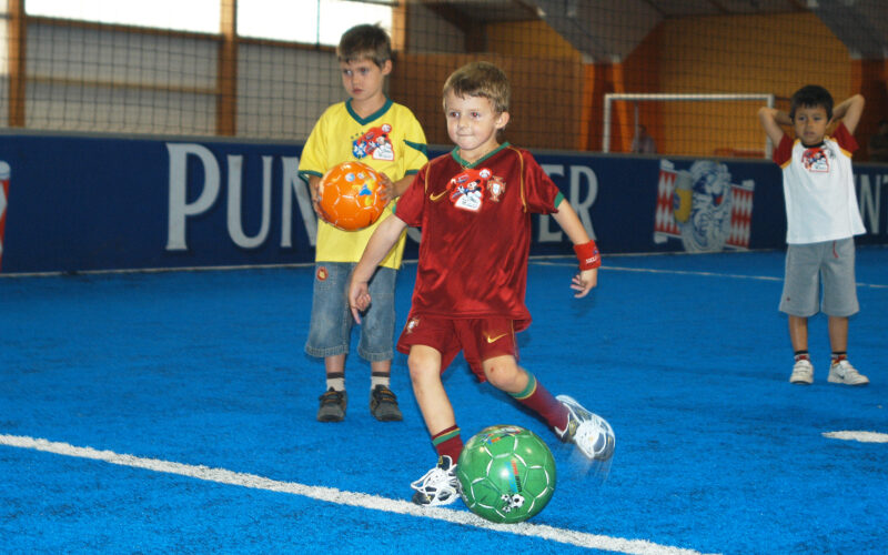 Drei Jungen stehen auf einem blauen Hallenfußballfeld, einer davon in einem roten Trikot, der einen grünen Fußball kickt, während die anderen zusehen.