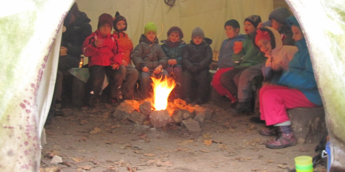 Eine Gruppe von Kindern mit Jacken und Hüten sitzt in einem Kreis um ein Lagerfeuer in einem Zelt, auf dem Boden liegen Blätter verstreut.