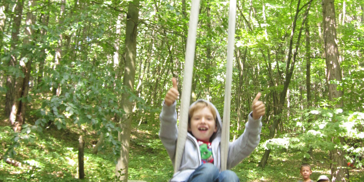 Ein Kind schaukelt lächelnd auf einer Seilschaukel in einem Wald, während zwei andere Kinder im Hintergrund zwischen den Bäumen stehen.
