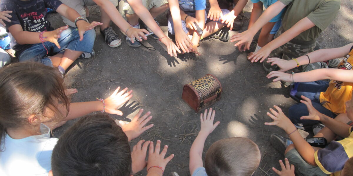 Eine Gruppe von Kindern sitzt in einem Kreis auf dem Boden und streckt ihre Hände nach einer kleinen Holzkiste in der Mitte aus.
