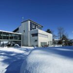 Modernes Gebäude mit großen Fenstern, umgeben von Schnee, Bäumen und einem klaren blauen Himmel, mit einem kleinen Gebäude im Hintergrund.