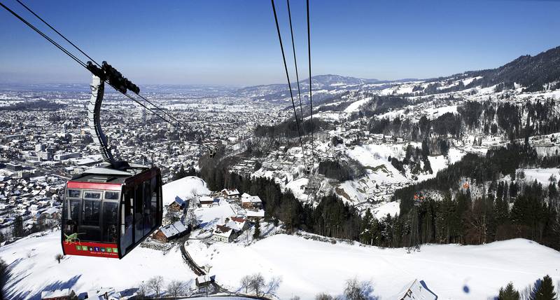 Eine rote Seilbahn fährt über eine verschneite Berglandschaft mit verstreuten Häusern und einer in der Ferne sichtbaren Stadt unter einem klaren blauen Himmel.