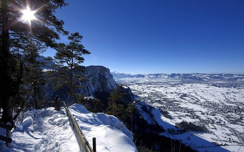 Schneebedeckter Aussichtspunkt auf einen Berg mit Bäumen auf der linken Seite, einem Holzgeländer und einem breiten Tal unter einem klaren blauen Himmel mit hellem Sonnenlicht.