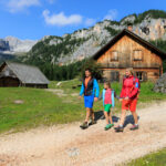 Drei Personen gehen auf einem Schotterweg in der Nähe von Holzhütten in einer Berglandschaft mit grünem Gras, Bäumen und felsigen Hügeln unter blauem Himmel.