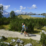 Zwei Personen wandern auf einem Schotterweg durch grüne Büsche mit Bergen, einem See und einem klaren blauen Himmel im Hintergrund.