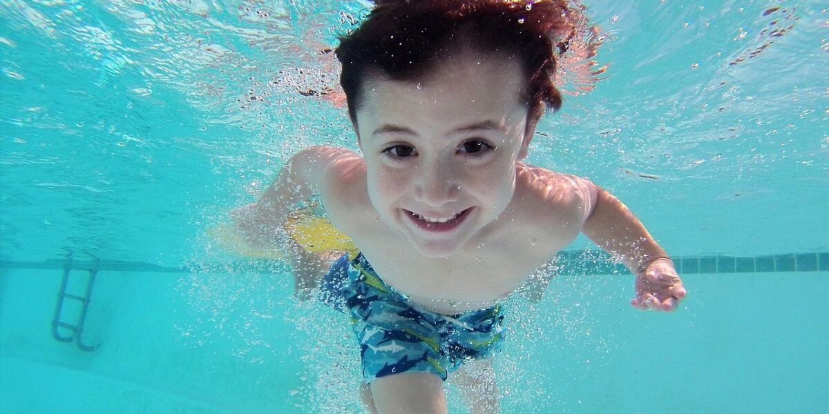 Ein Junge in blauer Badehose schwimmt in einem Schwimmbecken unter Wasser und lächelt in die Kamera. Im Hintergrund ist die Badeleiter zu sehen.