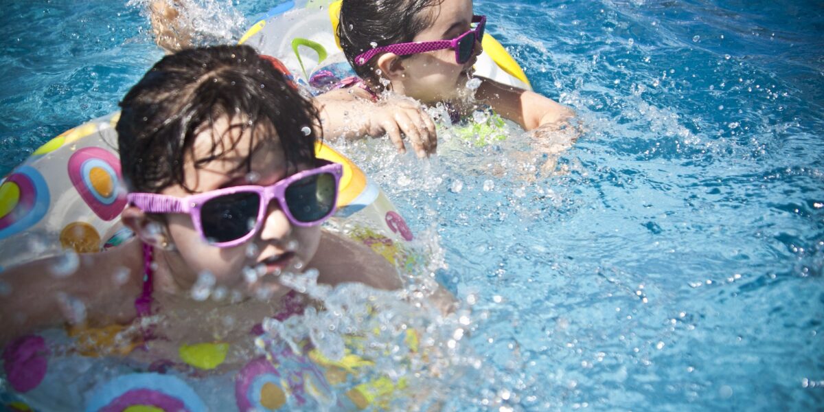 Zwei Kinder, die Sonnenbrillen und bunte Schwimmringe tragen, spielen in einem Schwimmbad, planschen im Wasser und genießen den sonnigen Tag.