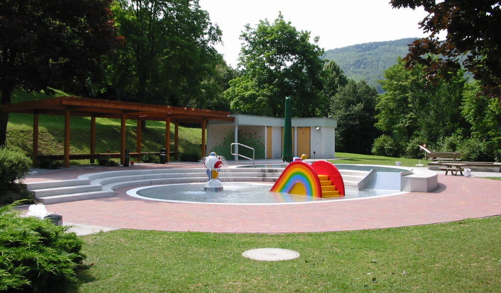 Ein kleines Kinderplanschbecken mit Tierfiguren und einer Regenbogenrutsche, umgeben von Gras, Bäumen, Bänken und einer hölzernen Schattenspenderstruktur.