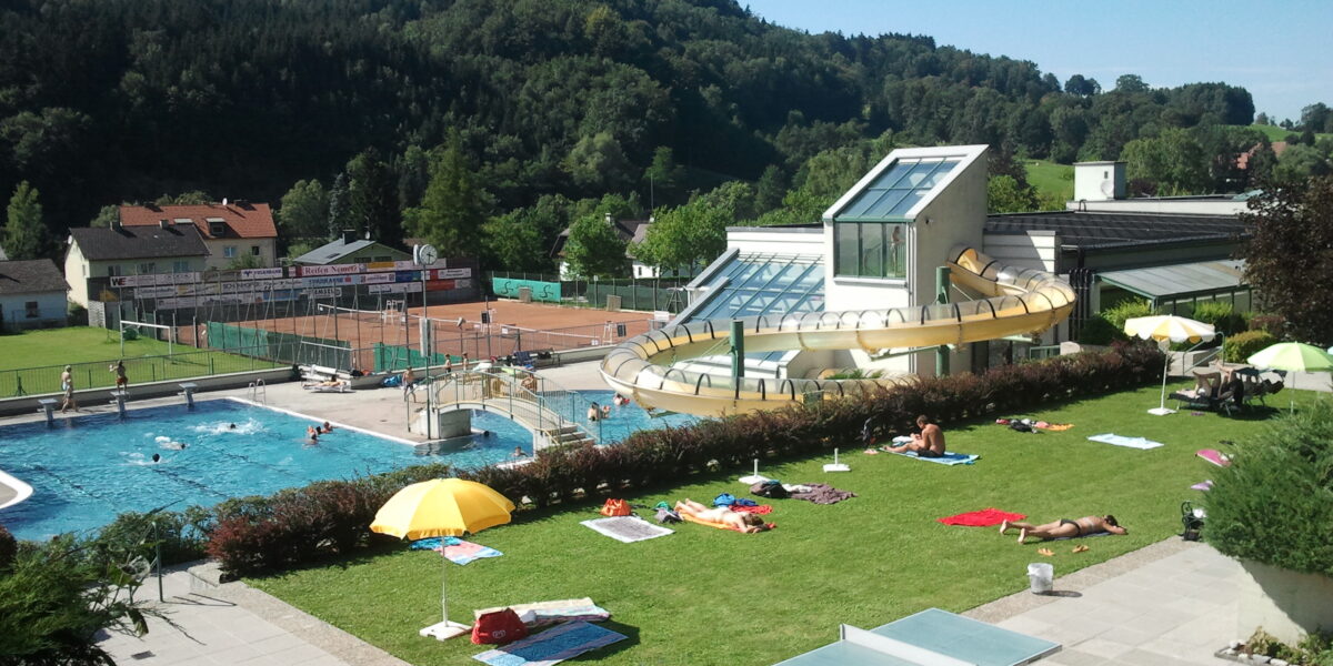 Freibadkomplex mit Wasserrutsche, Liegestühlen auf der Wiese, Tennisplätzen und einer malerischen Kulisse aus grünen Hügeln und Bäumen unter einem strahlend blauen Himmel.