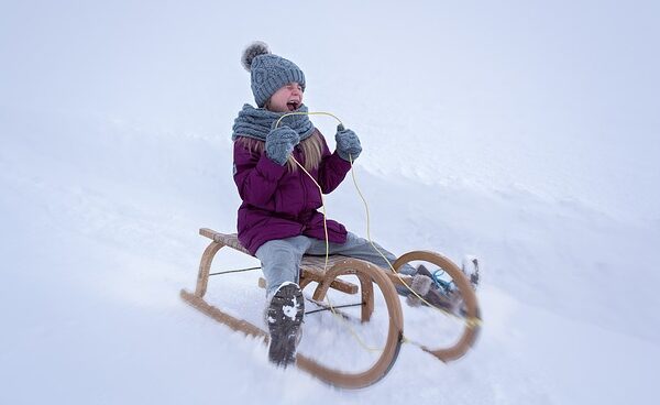Ein winterlich gekleidetes Kind fährt auf einem Holzschlitten einen verschneiten Hügel hinunter, wobei es sich an einem Seil festhält und einen fröhlichen Gesichtsausdruck macht.