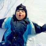 Ein Kind mit blauem Wintermantel und Mütze liegt mit ausgestreckten Armen im Schnee und macht ein Gesicht, als würde es ausatmen oder überrascht sein.