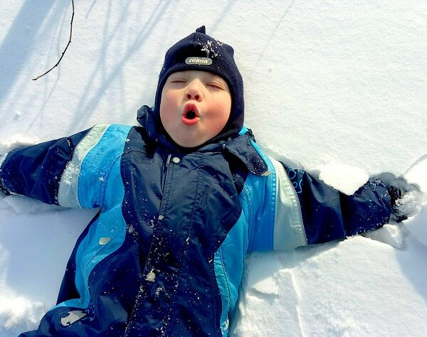 Ein Kind mit blauem Wintermantel und Mütze liegt mit ausgestreckten Armen im Schnee und macht ein Gesicht, als würde es ausatmen oder überrascht sein.