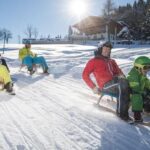 Vier Personen rodeln auf einer verschneiten Piste im hellen Wintersonnenlicht bergab, mit einem Holzhaus und Bäumen im Hintergrund.