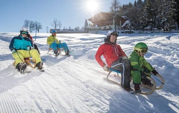 Vier Personen rodeln auf einer verschneiten Piste im hellen Wintersonnenlicht bergab, mit einem Holzhaus und Bäumen im Hintergrund.