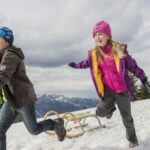 Zwei Kinder in Winterkleidung laufen und ziehen Holzschlitten auf einem verschneiten Hügel mit Bergen und einer Hütte im Hintergrund.
