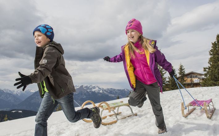 Zwei Kinder in Winterkleidung laufen und ziehen Holzschlitten auf einem verschneiten Hügel mit Bergen und einer Hütte im Hintergrund.