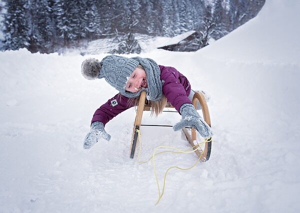 Eine Person in Winterkleidung fährt auf einem Holzschlitten einen verschneiten Hügel hinunter, wobei sie sich mit behandschuhten Händen nach vorne streckt. Im Hintergrund sind schneebedeckte Bäume zu sehen.