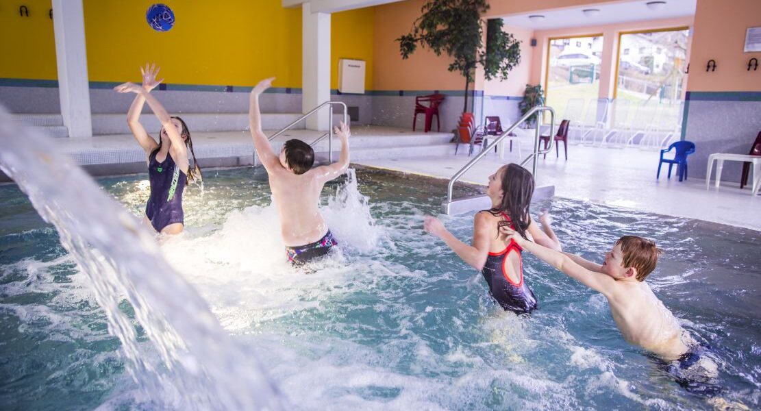 Vier Kinder spielen zusammen und planschen in einem Hallenbad mit heller Beleuchtung und bunten Wänden.