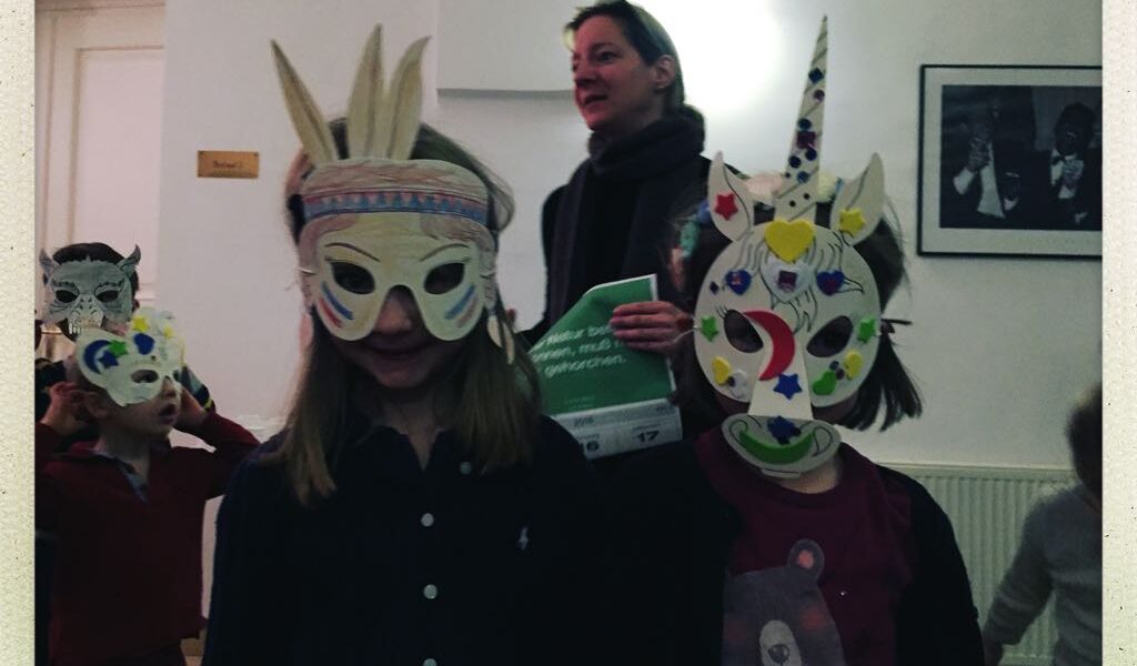 Vier Kinder mit bunten, handgefertigten Tier- und Einhornmasken stehen in einem Haus, während ein Erwachsener im Hintergrund Papiere hält.