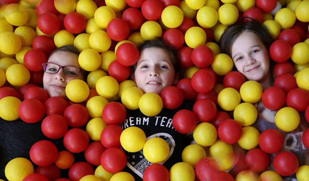 Drei Kinder liegen in einer mit gelben und roten Plastikbällen gefüllten Ballgrube, wobei nur ihre Gesichter und ein Teil ihres Oberkörpers zu sehen sind.