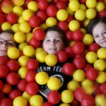 Drei Kinder liegen in einer mit gelben und roten Plastikbällen gefüllten Ballgrube, wobei nur ihre Gesichter und ein Teil ihres Oberkörpers zu sehen sind.
