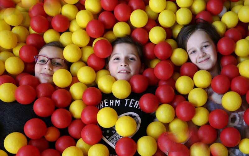 Drei Kinder liegen in einer mit gelben und roten Plastikbällen gefüllten Ballgrube, wobei nur ihre Gesichter und ein Teil ihres Oberkörpers zu sehen sind.