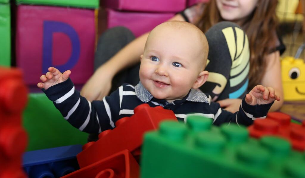 Ein Baby in einem gestreiften Hemd sitzt lächelnd zwischen großen bunten Spielzeugblöcken, im Hintergrund ein älteres Kind.