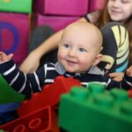 Ein Baby in einem gestreiften Hemd sitzt lächelnd zwischen großen bunten Spielzeugblöcken, im Hintergrund ein älteres Kind.