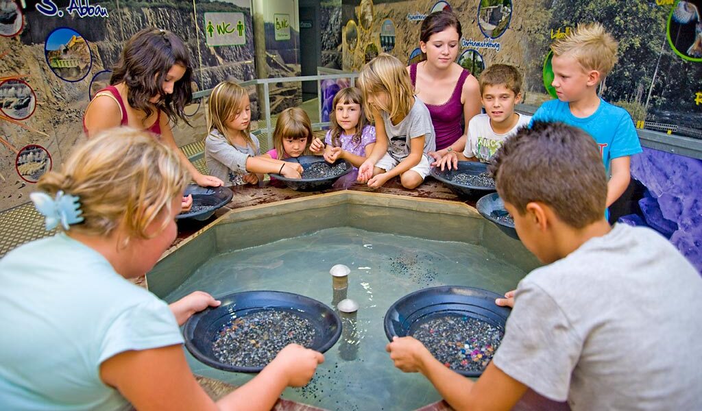 Eine Gruppe von Kindern und ein Erwachsener versammeln sich um einen kreisförmigen Wassertisch und suchen nach Steinen oder Mineralien in einer pädagogischen Umgebung.