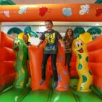 Drei Kinder spielen und hüpfen auf einer bunten aufblasbaren Struktur, die mit Cartoon-Insekten und Pflanzen verziert ist.
