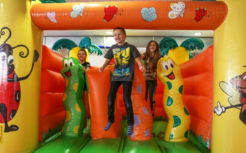 Drei Kinder spielen und hüpfen auf einer bunten aufblasbaren Struktur, die mit Cartoon-Insekten und Pflanzen verziert ist.