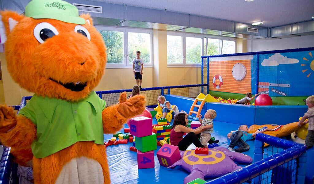 Eine Person in einem orangefarbenen Eichhörnchen-Maskottchenkostüm steht in der Nähe von Kindern und Erwachsenen, die mit großen Blöcken und Spielzeug in einem Indoor-Spielbereich spielen.
