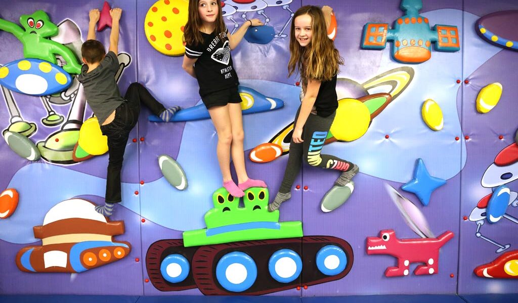 Drei Kinder klettern und posieren auf einer farbenfrohen Spielwand, die mit Planeten, Raumschiffen und Zeichentrickfiguren dekoriert ist.