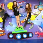Drei Kinder klettern und posieren auf einer farbenfrohen Spielwand, die mit Planeten, Raumschiffen und Zeichentrickfiguren dekoriert ist.