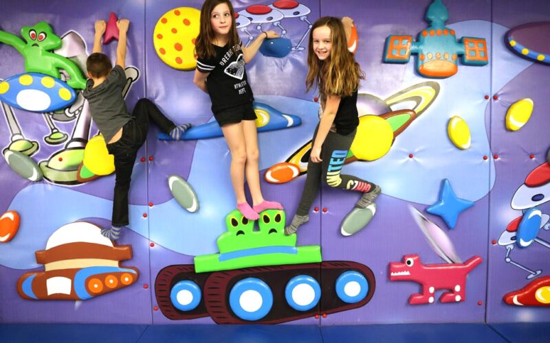 Drei Kinder klettern und posieren auf einer farbenfrohen Spielwand, die mit Planeten, Raumschiffen und Zeichentrickfiguren dekoriert ist.